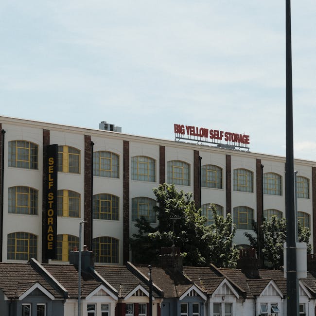 A multi-storey commercial building with a white facade and large, yellow-framed windows, featuring a prominent red sign on the rooftop reading 'BIG YELLOW SELF STORAGE'. Attached to the building is a vertical black sign with yellow text stating 'SELF STORAGE'. Below, the building is situated behind a row of small residential houses with tiled roofs and chimneys, which are partially obscured by trees and shrubs. In the foreground, a lamp post and a streetlight are visible, with the sky above showing a partly cloudy day. The scene suggests a typical urban environment where house removals or furniture transport might involve navigating around nearby structures during a home relocation or moving process facilitated by companies like Man and Van New Cross.