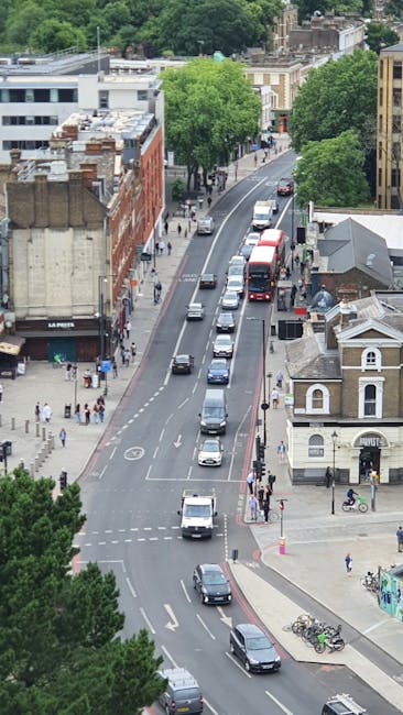 An aerial view of a busy residential street during daytime in New Cross, showing parked cars along both sides of the road, a line of moving vehicles including a red bus and several smaller cars, and pedestrians walking on the sidewalks. There are trees lining the street providing greenery, alongside a mix of commercial and residential buildings with brick facades and flat roofs. The scene captures typical urban traffic and foot traffic, illustrating a common access challenge for house removals and furniture transport in the area. The image reflects the environment where Man and Van New Cross offers house removals services, highlighting potential obstacles during loading and unloading processes at accessible street locations.