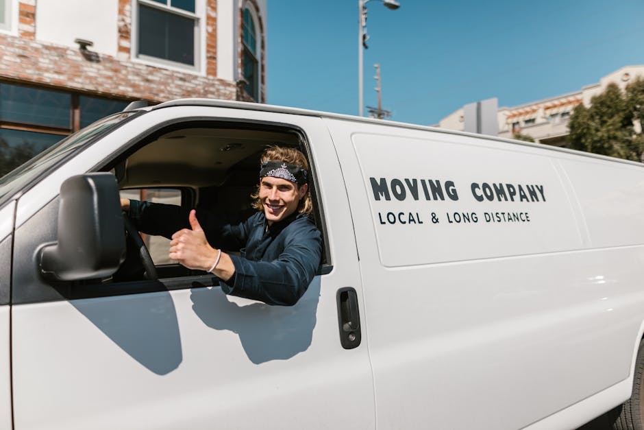 A smiling man with long, wavy hair, wearing a black headband and dark clothing, is sitting in the driver's seat of a white moving van with the door open. He is giving a thumbs-up gesture and looking out towards the camera. The van displays the company name 'Moving Company' along with the service description 'Local & Long Distance' on its side. The vehicle is parked outdoors on a street, with a residential building, trees, and a blue sky visible in the background. Inside the van, visible behind the man, are packed cardboard boxes and some furniture wrapped in protective materials, indicating a home relocation or furniture transport process. The scene captures an active loading or unloading moment, typical of house removals and moving services. Man and Van New Cross, specialising in removals, is identifiable through this visual context, aligning with content on common access problems for New Cross removals vans.