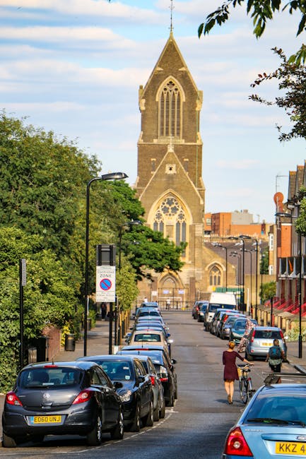 A red moving truck with the branding 'ALCIDES' on its sign, positioned on a city street in front of multi-story residential buildings with balconies, some with plants. The truck is parked partially in
