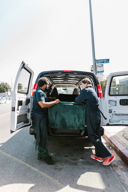 Two movers from Man and Van New Cross are engaged in a house removal process, carefully loading a large, heavy piece of furniture covered in a dark green protective blanket into the back of a white van. The van's rear doors are wide open, revealing a spacious interior prepared for furniture transport. One mover, dressed in dark clothing, is positioned on the left side of the image, lifting or stabilising the furniture, while the other, wearing a darker jacket, red sports shoes, and a cap, is on the right, assisting with the placement and ensuring proper handling. The scene takes place outdoors on a paved street near a lamppost with a blue street sign labeled 'Somerset.' Bright daylight highlights the environment, indicating a daytime house relocation or packing and moving operation, with additional context suggesting the transport of household items for students leaving halls, as referenced in the Goldsmiths removals service for New Cross. Man and Van New Cross provides specialised removal services supported by such loading and furniture transport activities.