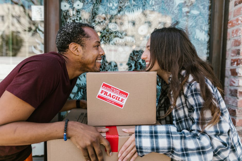 A man and a woman are engaged in a home relocation activity outdoors, with both holding cardboard boxes. The man, on the left, has short dark curly hair, wearing a maroon T-shirt and a blue beaded bracelet, and is smiling while facing the woman. The woman, on the right, has long brown hair, dressed in a blue and white plaid shirt, and is smiling back at the man. They are standing close to each other, each holding the sides of a medium-sized cardboard box. On top of this box, there is a smaller box with a red and white label that reads “Fragile” and “Handle with care,” indicating it contains delicate items. The background features a glass window with decorative illustrations and a brick wall, suggesting the scene occurs outside a residential property. The image visually captures a moment during the packing and furniture transport process involved in a house removal, illustrating careful handling and teamwork in moving logistics, supported by professional removals services like those provided by Man and Van New Cross.