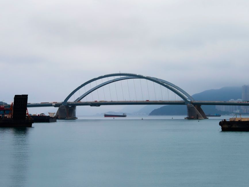 A large modern arch bridge spans across a calm body of water, with a boat sailing underneath. The bridge features a prominent curved arch with suspension cables supporting the roadway above. The scene is set during overcast weather, with grey skies and a faint outline of distant hills or mountains in the background. In the foreground, there are part of a dock or industrial area with dark structures and mooring points. The water reflects the bridge and sky, creating a tranquil atmosphere. This image relates to logistics and transport, similar to house removals and furniture transport services provided by Man and Van New Cross, emphasizing the importance of careful handling and planning during home relocation or moving processes.