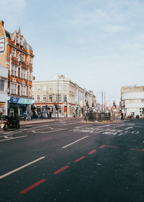 A wide view of a busy street scene in New Cross, Lewisham, featuring multi-storey Victorian-style buildings with decorative facades, some with bay windows, and ground-floor retail shops. The sky is partly cloudy with natural daylight illuminating the area. On the pavement, several pedestrians are walking, and a few are standing near shop entrances. A black motorcycle is parked on the side of the road near a designated parking zone. In the foreground, an intersection with road markings including a red dashed line indicating a no-parking zone and directional arrows are visible. The street appears to be part of a home relocation or furniture transport process, possibly managed by Man and Van New Cross, with no visible moving equipment, furniture, or boxes in this particular image but representing an urban environment suitable for house removals and professional moving services.