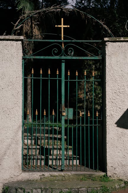 A green metal gate with decorative scrollwork and gold finials at the top, set between two white concrete pillars. The gate features vertical bars and a padlock securing the locking mechanism, with a small latch visible. Behind the gate, stone steps lead upward into a garden area, partially shaded by surrounding trees, with dense foliage and natural light filtering through. A wooden bench is partly visible beyond the steps. The gate has a cross mounted on top, symbolizing religious significance, and the setting appears to be an outdoor entrance to a property. This scene represents a secure, traditional entrance that might be encountered in residential settings during a home relocation or furniture transport process, with the gate providing access to a private outdoor space. Man and Van New Cross facilitates moving services, including access through such gates during packing and loading activities.