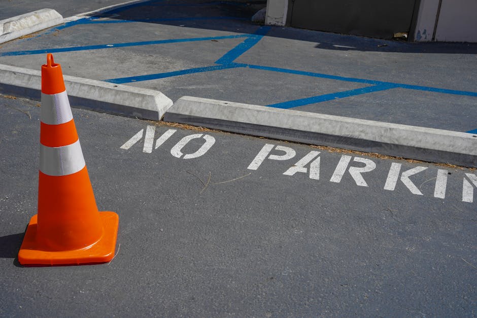 A bright orange traffic cone positioned on a paved parking area in front of a designated no parking zone, which is marked with white painted letters on the asphalt reading 'NO PARKING.' Behind the cone, there are concrete wheel stops and blue painted lines indicating parking bays, with some of these lines slightly faded. To the right, a grey building wall or gate is partially visible. The scene is outdoors under natural daylight, with shadows cast by the cone and other objects. This setting suggests an area where vehicle movement is restricted, likely relevant to house removals or moving logistics, and highlights the importance of adhering to parking restrictions during a home relocation, as managed by [COMPANY_NAME].