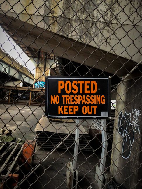 A close-up view of a metal wire fence with a black and orange sign attached that reads 'POSTED NO TRESPASSING KEEP OUT'. The fence is situated in front of a construction or industrial site, with concrete structures, graffiti on the wall to the right, and construction materials visible behind the fence. Inside the enclosed area, there are wooden pallets, a small orange container, and some boxes, indicating storage or ongoing work. The environment appears under a bridge or overpass, with concrete beams and supports overhead, and the lighting is natural, suggesting daytime. The scene reflects a restricted area, typical of property safety signage, relevant to house removals and relocation logistics companies like Man and Van New Cross, especially in contexts involving moving restrictions or site security during home relocation processes.