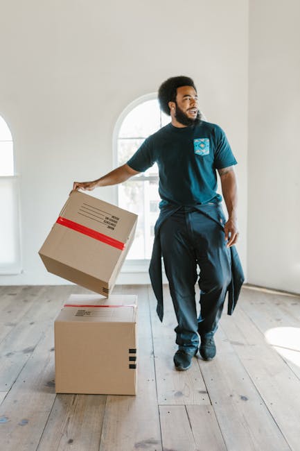 A man with a full beard and dark curly hair, wearing a navy blue t-shirt with a logo on the chest and dark trousers, stands inside a spacious, well-lit room with wooden flooring and curved windows. He is holding a cardboard moving box in his right hand while standing next to two additional boxes on the floor, one with red tape and the other unsealed, indicating an ongoing home relocation process. The room appears empty aside from the boxes, and the natural light from the windows highlights the clean, minimal environment. This scene depicts the packing and loading phase of furniture transport and packing for a house move, illustrating the activity of a professional removals service such as Man and Van New Cross.