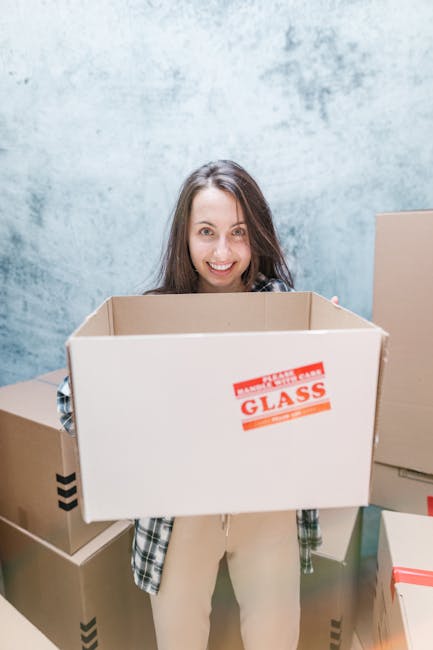 A woman with long brown hair and a friendly expression is standing indoors against a light, textured wall, holding an empty medium-sized cardboard box labeled 'Fragile Glass' with red printing. She is dressed in casual clothing, including a plaid shirt and light-colored trousers. Behind her, there are several other packed cardboard boxes of various sizes, some sealed with tape and others partially open, stacked on the floor. The scene depicts the packing stage of a house move, with packaging materials such as cardboard boxes and protective wrapping visible, and a focus on the preparation for a home relocation or furniture transport. The environment is well-lit, emphasizing a professional and organized approach to moving services, as provided by Man and Van New Cross, supporting efficient and careful packing and loading processes for an urgent same-day house removal.
