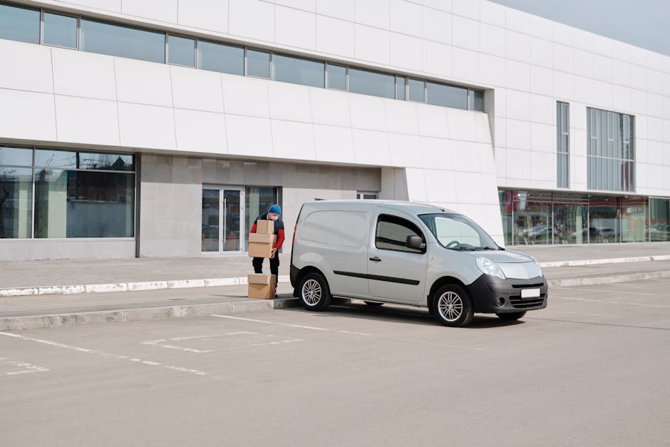 A man wearing a red jacket and blue beanie is standing on a paved parking area outside a modern white building, holding three cardboard boxes of varying sizes, preparing to load them into a grey commercial van parked nearby. The van is positioned close to the building's entrance, with the driver’s side facing the camera, and appears to be in the process of home relocation or furniture transport. The background features large glass windows and an urban environment, with no other vehicles or pedestrians visible at the moment. The scene is captured during daylight under overcast skies, emphasizing a neutral, professional approach to moving services. The image illustrates a typical loading process, aligned with house removals, and demonstrates the use of trolleys or manual carrying of boxes for efficient packing and moving operations, as undertaken by companies like Man and Van New Cross, especially relevant to parking regulations and optimal loading spots in SE14 or New Cross areas.