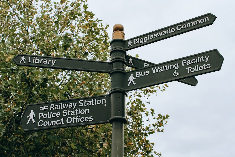 A black metal directional signpost with multiple rectangular signs attached, each pointing in different directions and labeled with white text for locations such as Biggleswade Common, Library, Railway Station, Police Station, Council Offices, Bus Waiting Facility, and Toilets. The signs feature pedestrian icons, with some signs including additional symbols like a wheelchair accessibility icon. The signpost stands outdoors against a backdrop of leafy green trees and an overcast sky, indicating a public area or neighbourhood environment observed during daylight. This setting may be relevant to home relocation or moving logistics, such as navigating to different local facilities during a house move, with Man and Van New Cross providing related removals and transport services.
