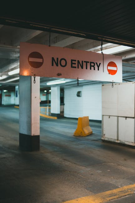 A white sign with black text reading 'NO ENTRY' and red circular symbols on either side, hanging from a dark ceiling above a parking garage. The concrete pillars and walls are painted in light and dark shades, with some yellow and black protective barriers on the floor, indicating a restricted area. In the background, the parking area features empty parking bays, with a smooth concrete surface and subtle lighting. This setting is part of a commercial or residential building, where moving companies such as Man and Van New Cross might coordinate logistics for home relocation or furniture transport. The image captures a section of a parking lot commonly used during the loading and unloading process, emphasizing controlled access for designated vehicles during house removals and other moving operations.